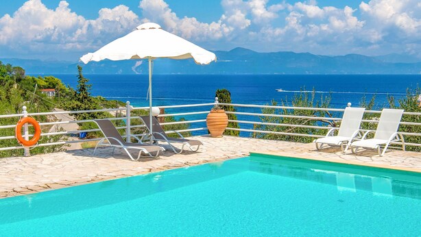 A pool area with lounge chairs, a white umbrella, and a view of the sea. - Dionisis Paxos Villa, 2-Story, Shared Pool, Sea Views, Modern Amenities (Yioliskari)