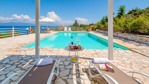Ein Poolbereich mit Liegestühlen, einem Tisch mit Früchten und einer Flasche sowie Blick auf das Meer.
