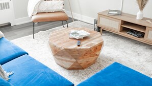 Un salon moderne avec une table basse hexagonale en bois, des canapés bleus, un tapis blanc et un buffet en bois sur lequel reposent une télécommande et un livre.