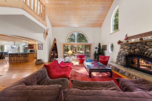 A spacious living room with a fireplace, red and brown sofas, and a large window overlooking a forest.