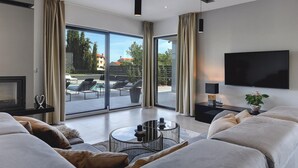 A modern living room with a large TV, comfortable seating, and a view of an outdoor pool area.