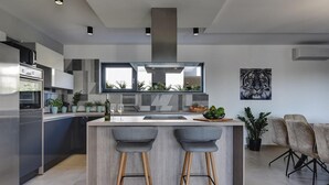 A modern kitchen with a central island, bar stools, built-in appliances, and a large window overlooking a garden.