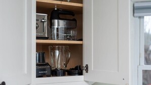Una cocina con un armario blanco que contiene una licuadora, un procesador de alimentos y una taza medidora. Hay un libro titulado 'The World in a Skill' sobre la encimera.