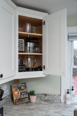 Una cocina con un armario blanco que contiene una licuadora, un procesador de alimentos y una taza medidora. Hay un libro titulado 'The World in a Skill' sobre la encimera.