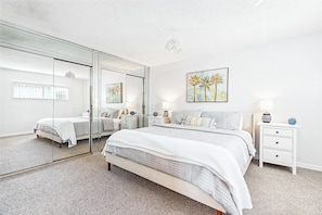 A bedroom with a large mirror, a bed with a quilted cover, bedside tables with lamps, and a wall painting. - Luxury Oceanfront Getaway on the Top Floor Penthouse Corner Unit Steps from the Beach (Cocoa Beach)