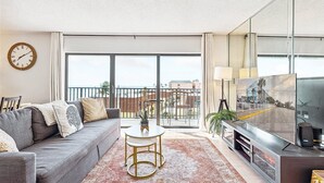 A modern living room with a grey sofa, a coffee table, and a TV. There is a balcony with a view of buildings and a railing.