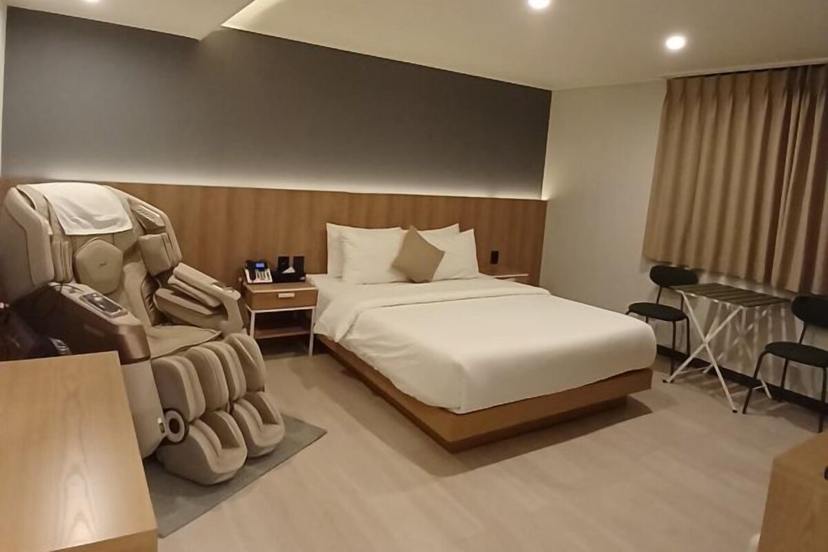A hotel room with a massage chair, a bed, a small table, and a window with curtains.