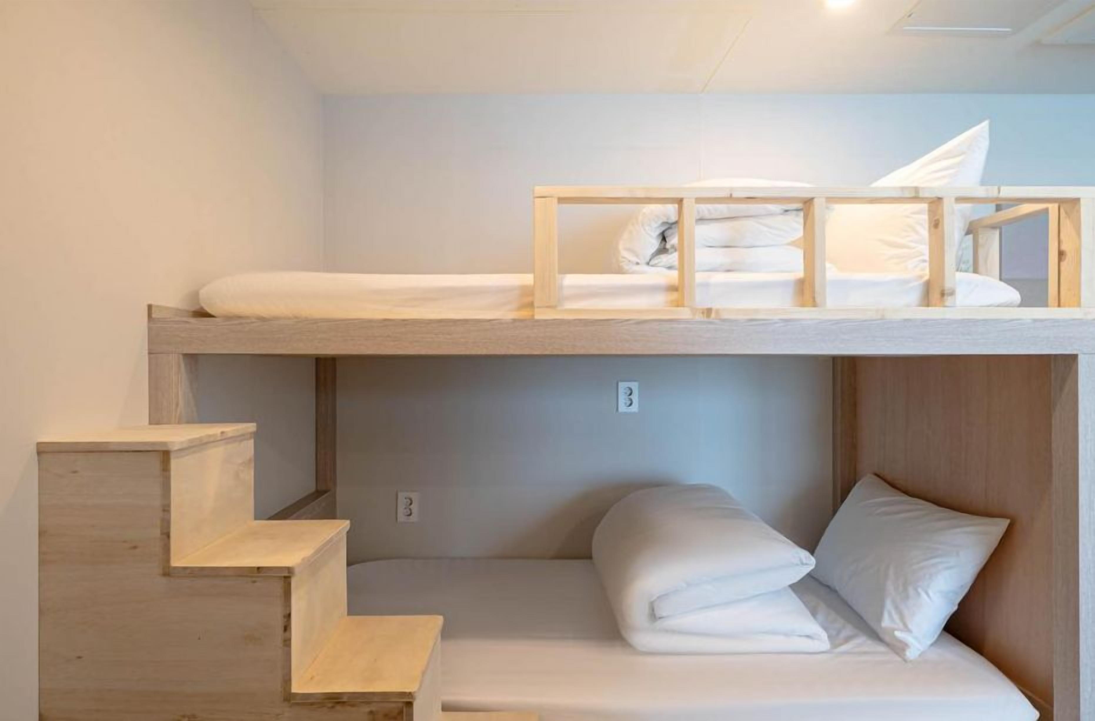 A bunk bed with a wooden frame and white bedding, a staircase leading to a lower bunk, and a wall-mounted light fixture.