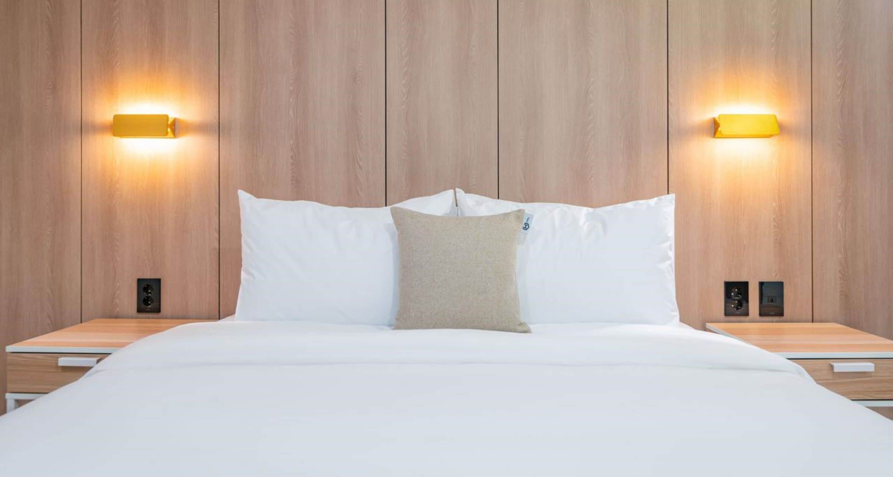 A hotel room with a wooden headboard, two bedside tables, a bed with white bedding, and two wall-mounted light fixtures.