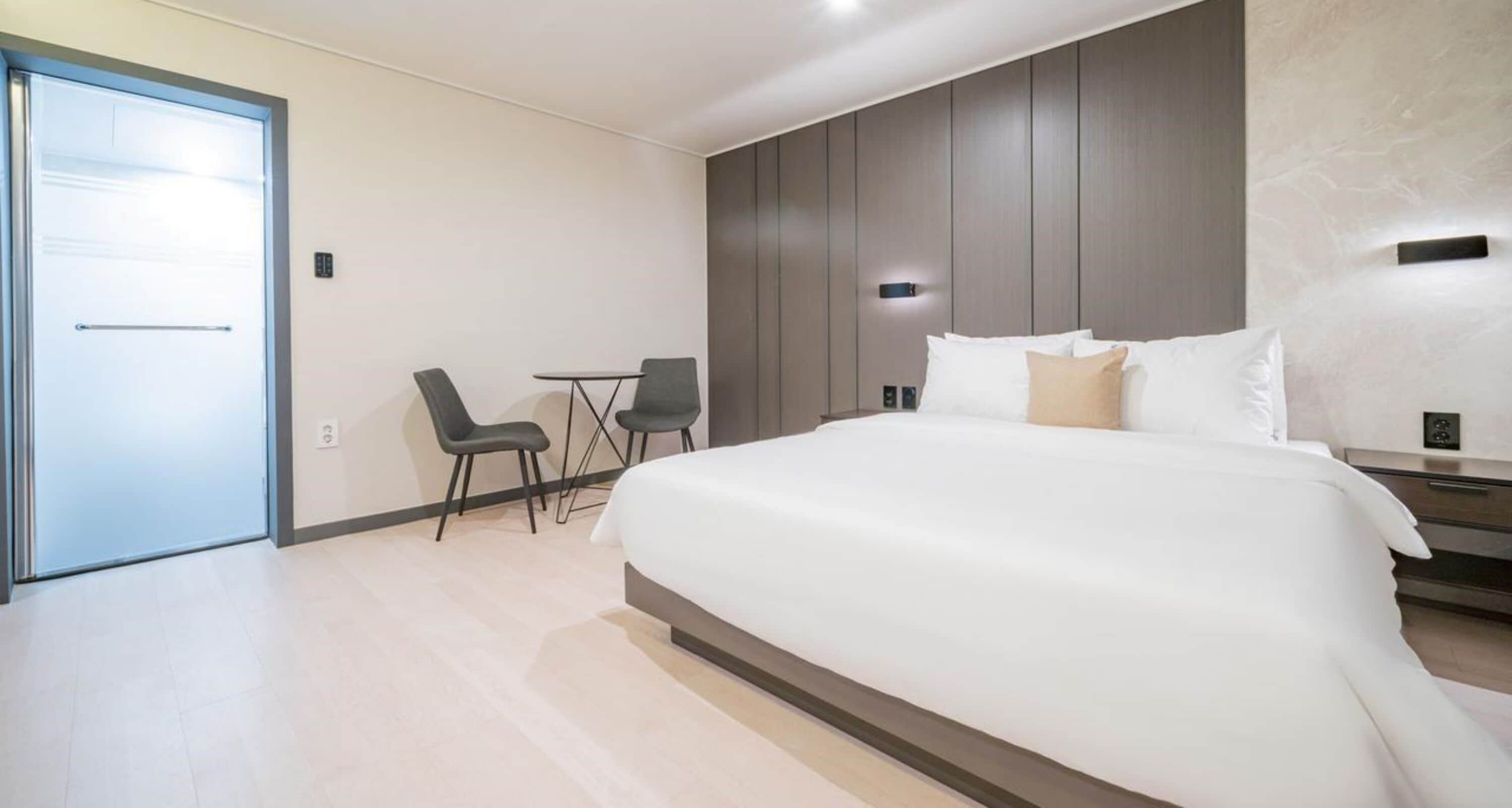 A modern hotel room with a large bed, a small desk with two chairs, and a door leading to a balcony.