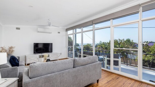 A modern living room with a large TV, grey sofa, and a balcony with a view of the city.