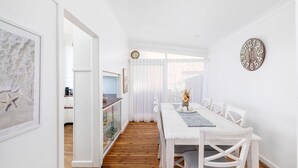 A bright, white dining area with a wooden table and chairs, a clock on the wall, and a view of a kitchen and living area through a glass door.