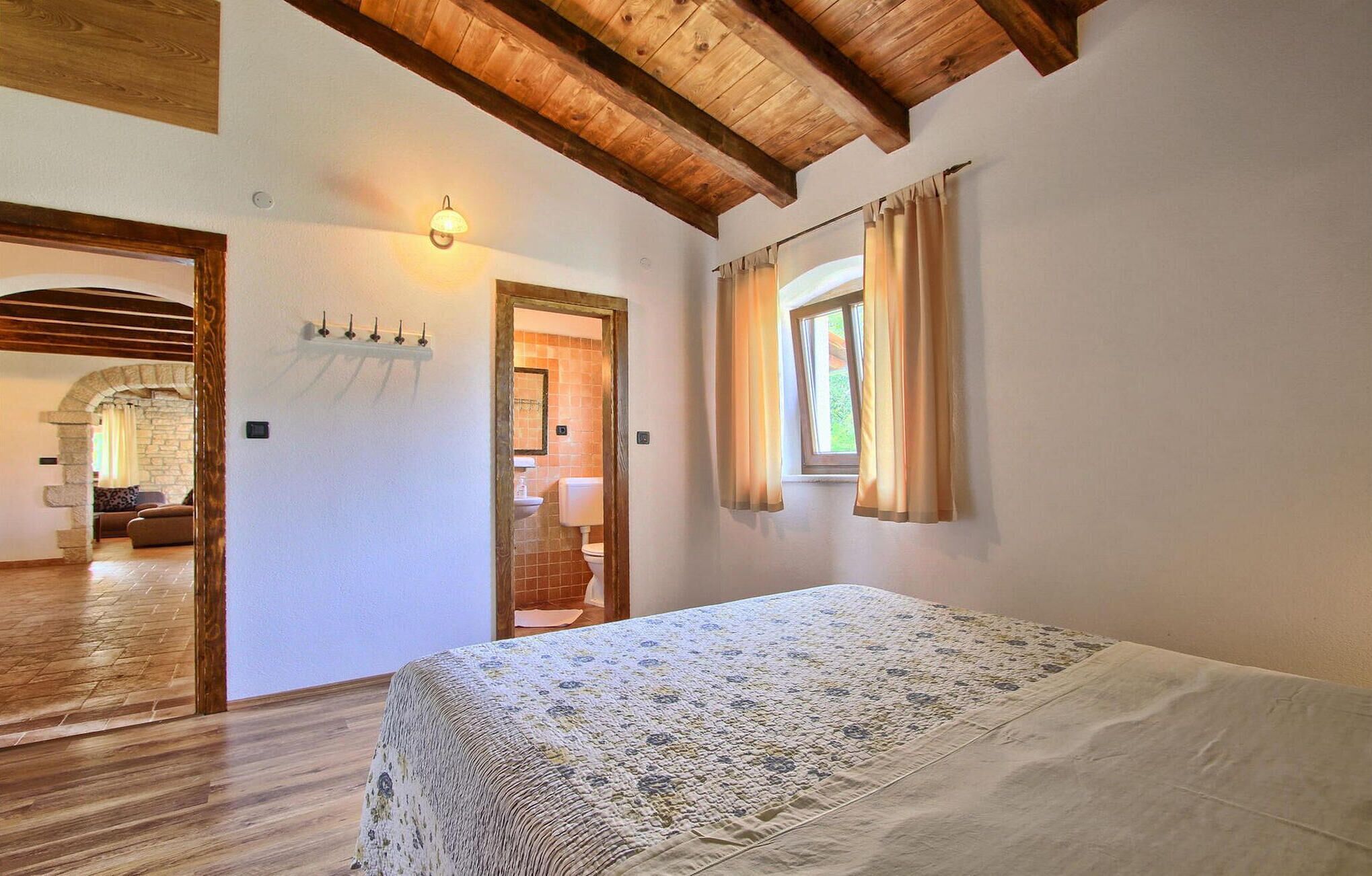 Un dormitorio con techo de madera, una cama con edredón estampado con motivos florales, una ventana con cortinas y un baño visible a través de un arco.