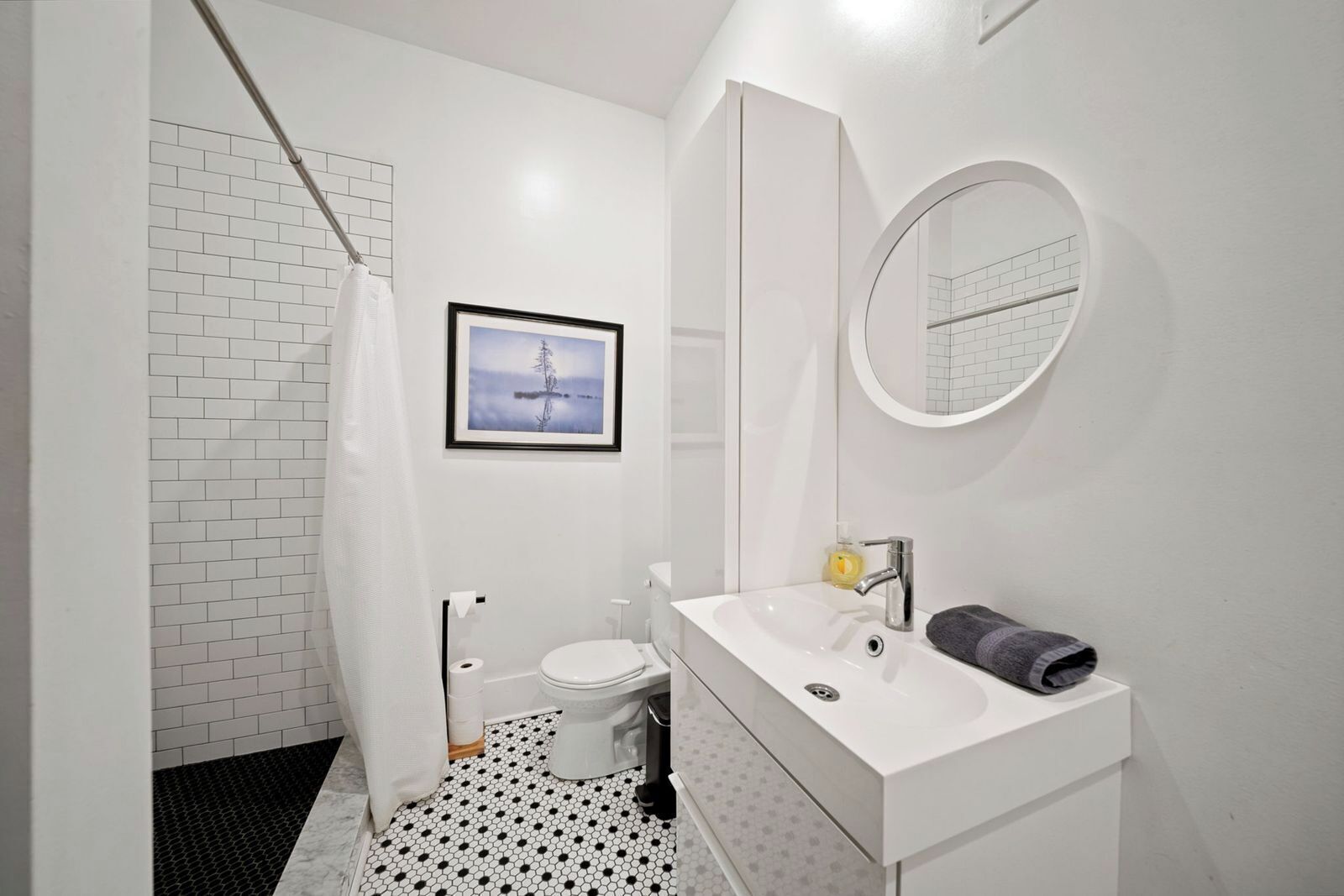 Une salle de bain avec un lavabo, des toilettes et une douche, un tableau encadré au mur et un miroir rond.
