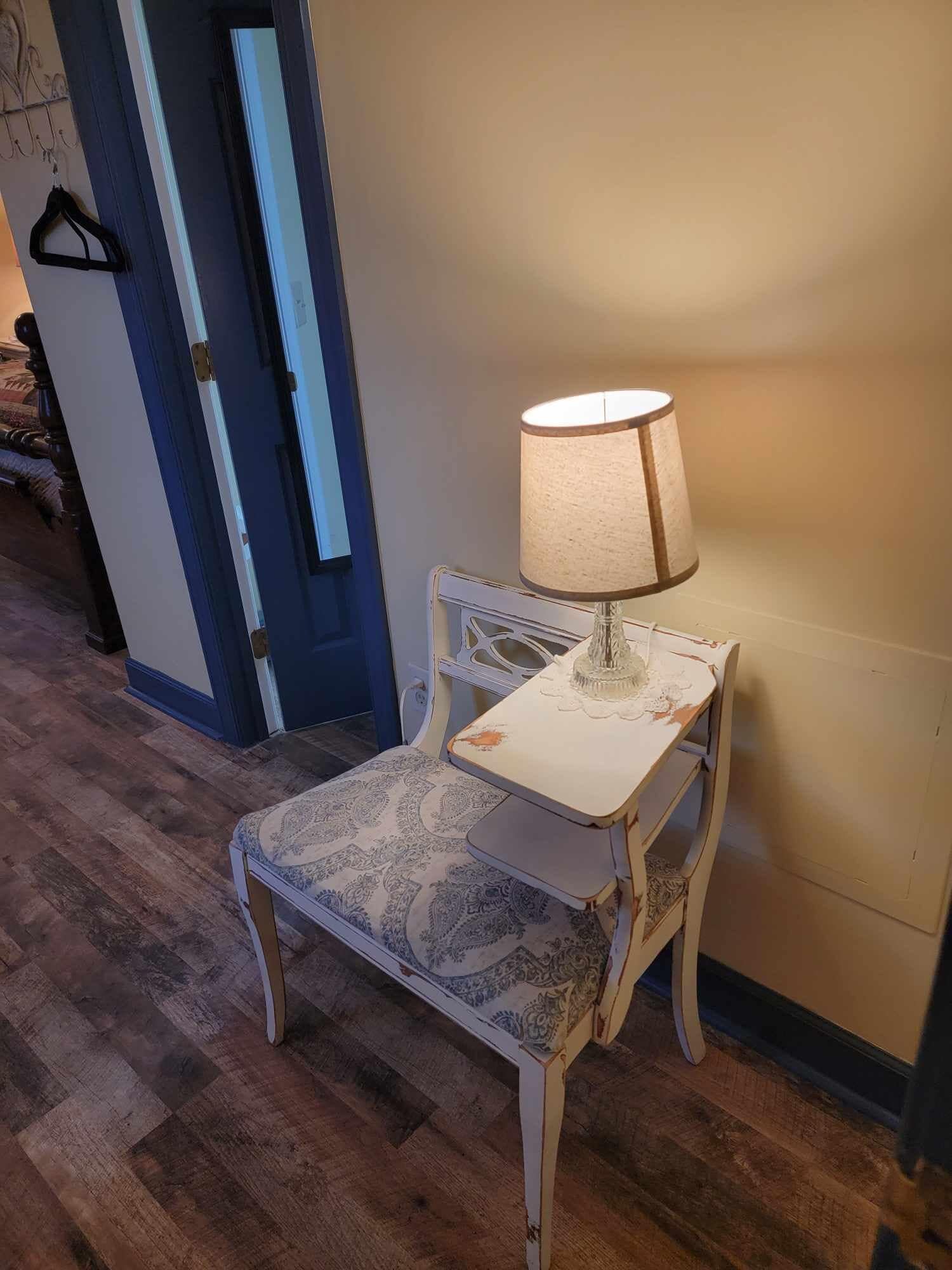 Ein kleiner, weißer Tisch im Vintage-Stil mit einer Tischdecke mit Muster und einer Lampe mit beiger Lampenschirm.