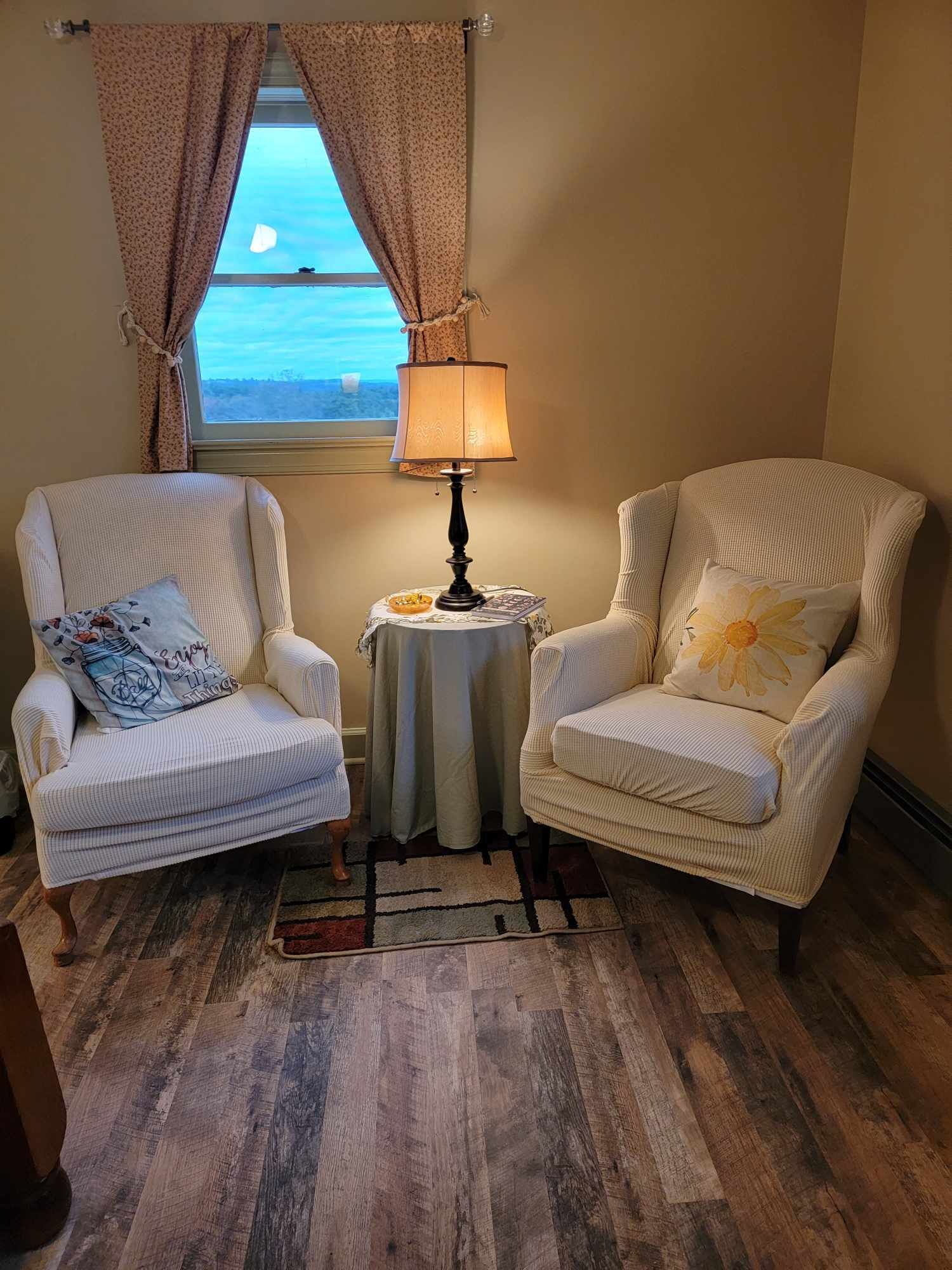 Una habitación acogedora con dos sillones, una mesita con lámpara y vistas al océano a través de una ventana con cortinas.