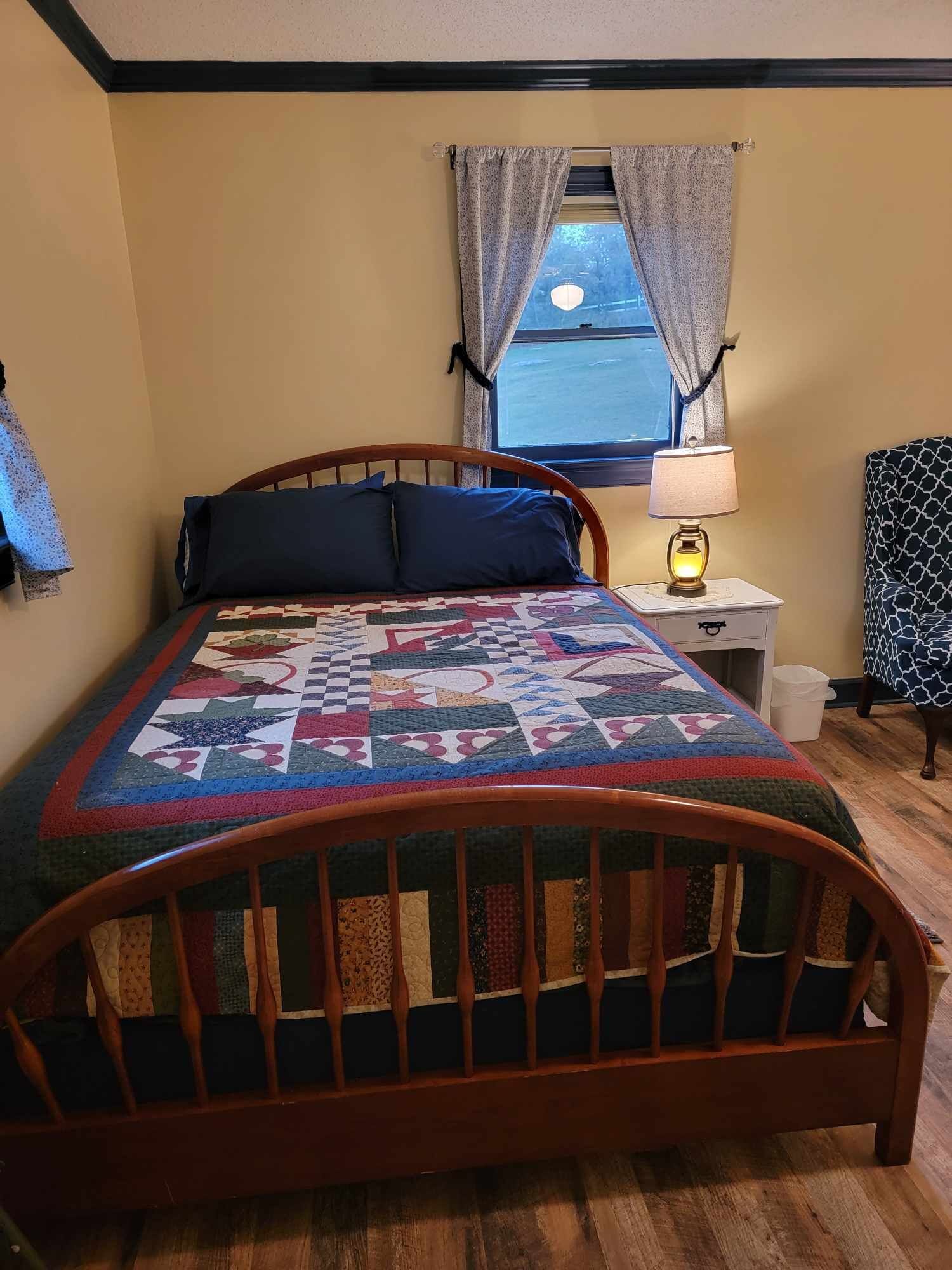 Ein Schlafzimmer mit einem hölzernen Bett, einer Steppdecke, einem Nachttisch mit Lampe, einem Sessel und einem Fenster mit Vorhängen.