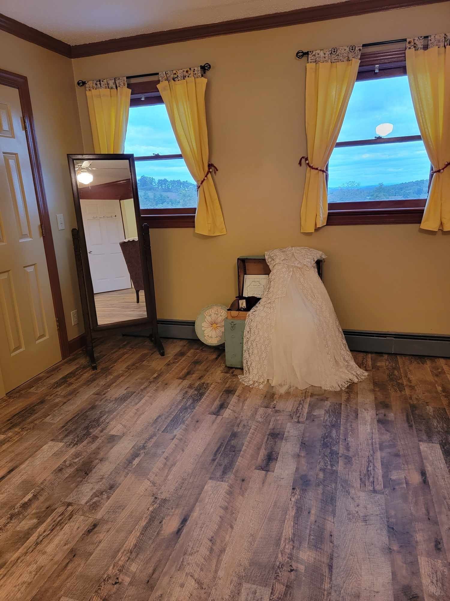 A room with wooden flooring, a full-length mirror, a chair, a small table, and a window with yellow curtains.