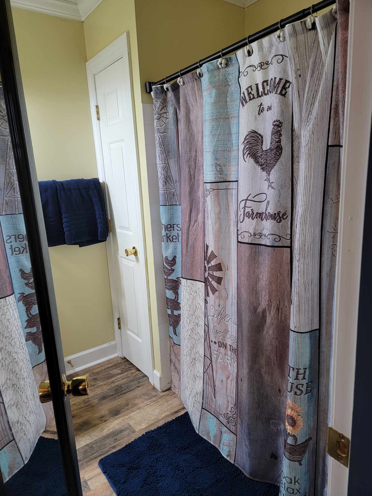 A bathroom with a farmhouse-themed shower curtain, a towel rack with a blue towel, and a wooden floor.