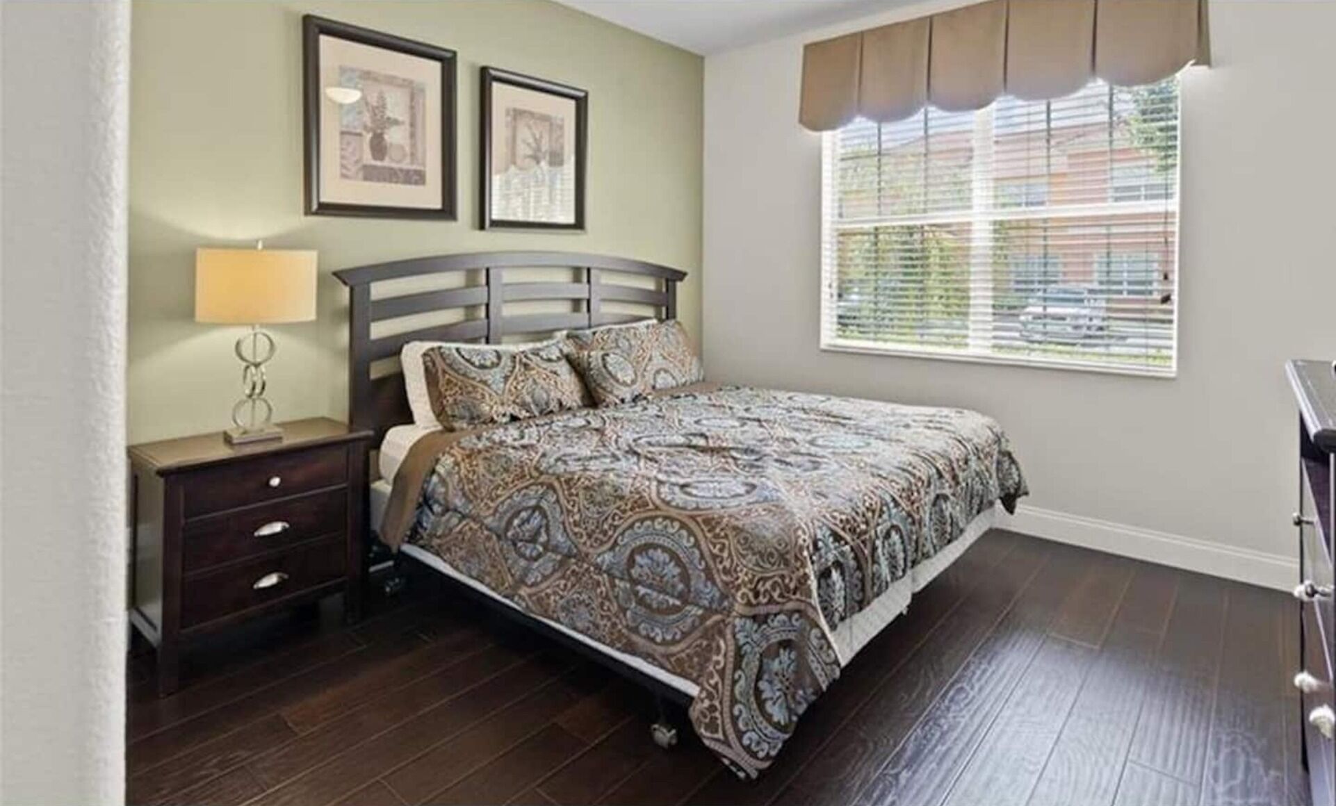A bedroom with a bed, a nightstand, a lamp, two framed pictures on the wall, and a window with blinds.
