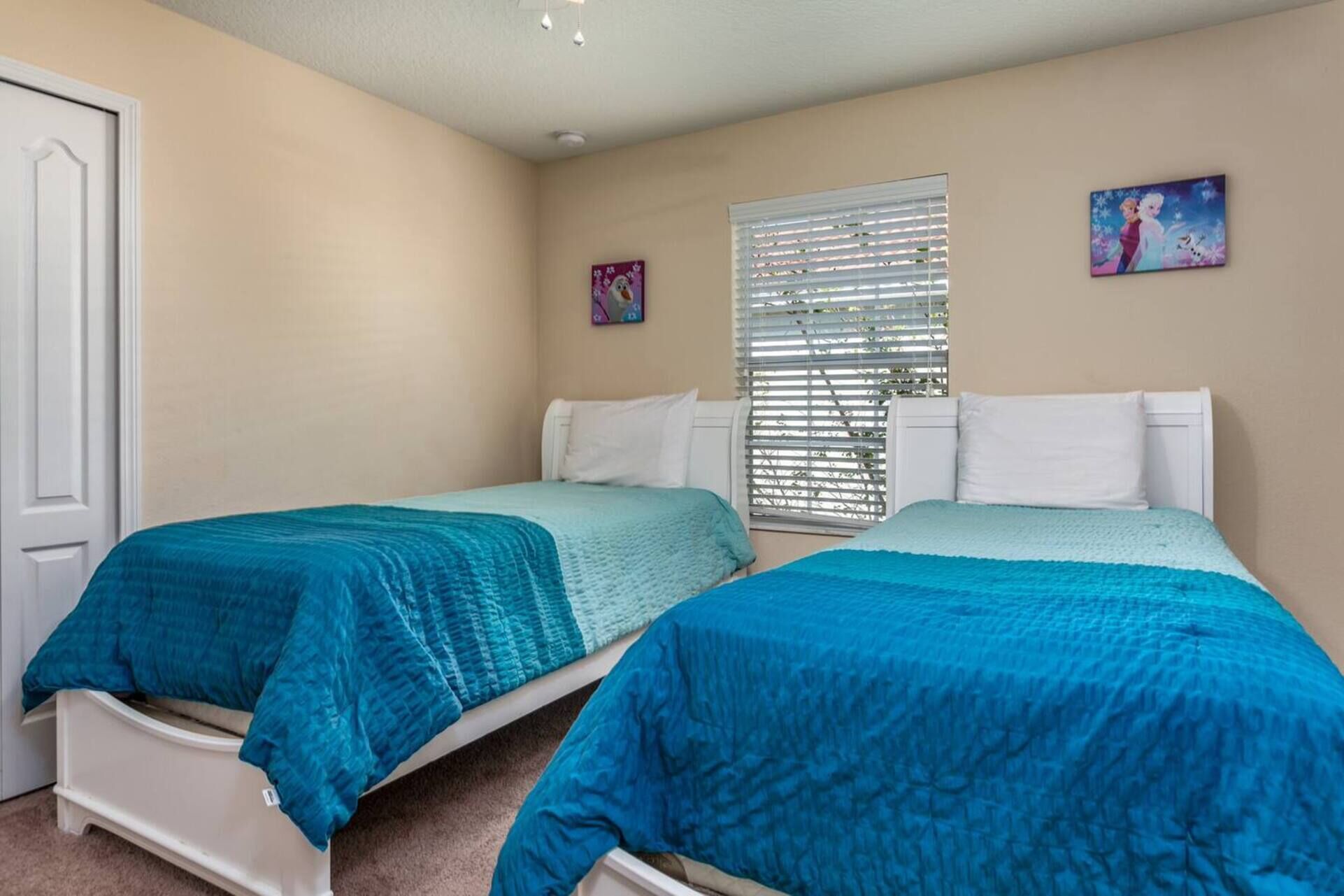 A bedroom with two single beds, beige walls, a window with blinds, and two small wall art pieces.