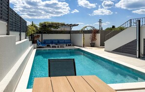 Une piscine avec une table en bois au premier plan, un ensemble de canapés bleus et une pergola ornée de plantes.