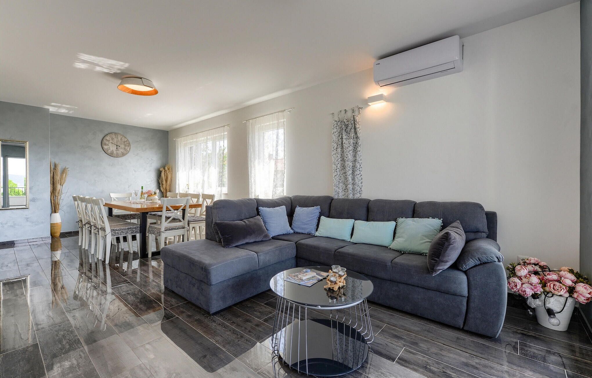 A modern living room with a grey sectional sofa, a round coffee table, and a dining area with a wooden table and white chairs.