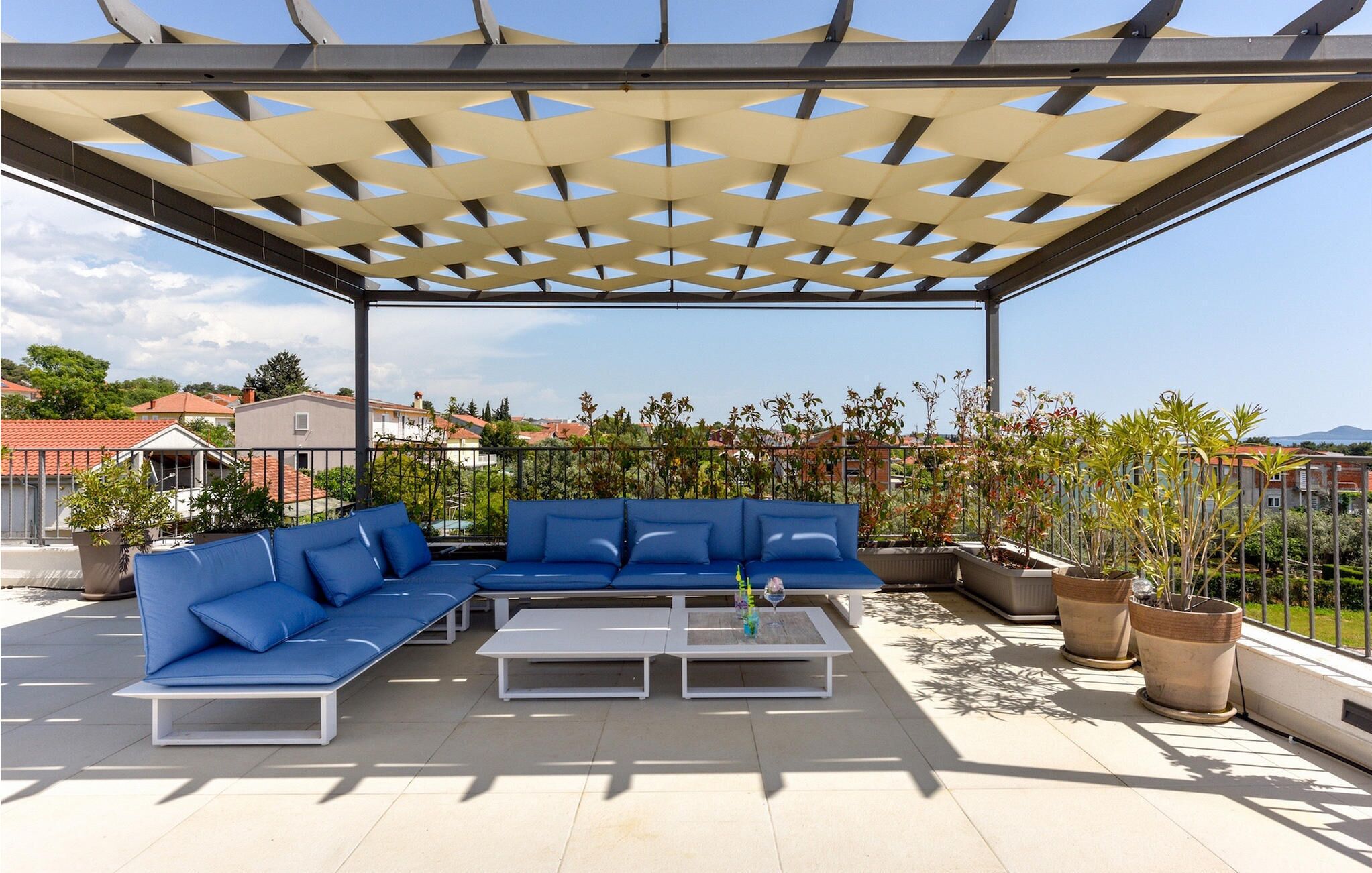 A covered outdoor seating area with a sectional sofa and two coffee tables, surrounded by potted plants.
