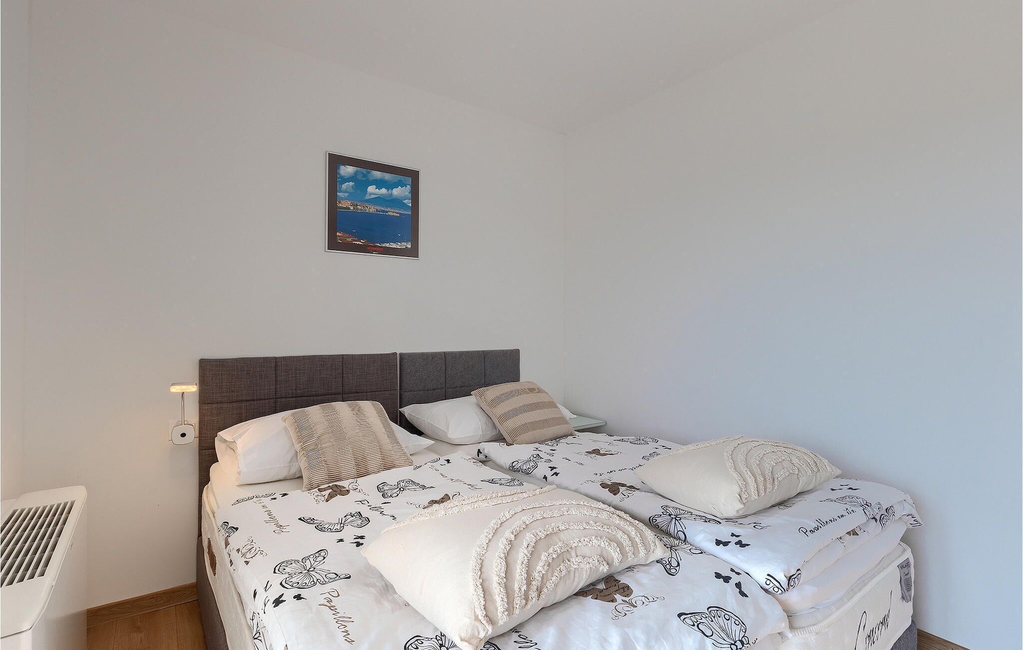 A bedroom with a bed, pillows, a nightstand, a wall-mounted lamp, and a framed picture.