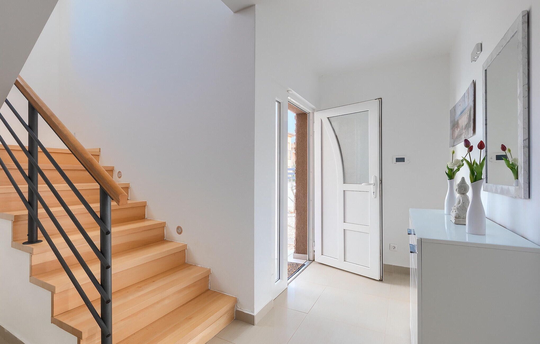 A modern interior with a wooden staircase, a white door leading to a balcony, and a white cabinet with a vase of flowers.