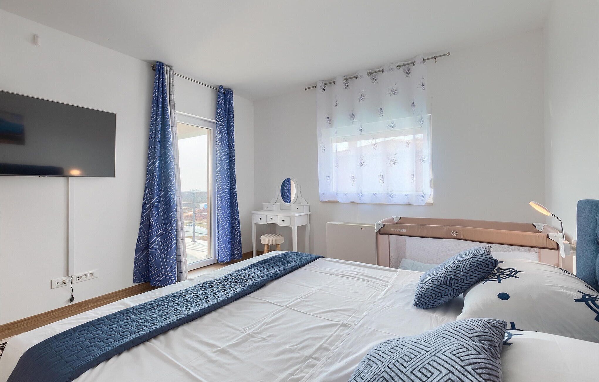 Een slaapkamer met een bed, een dressoir, een spiegel, een raam met gordijnen, en een aan de muur bevestigde televisie.