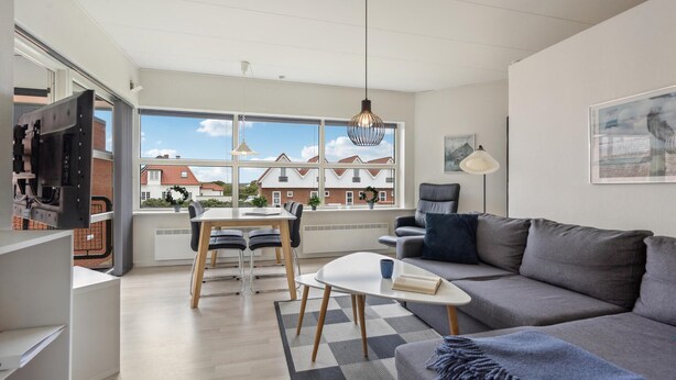 Un salon moderne avec un canapé gris, une petite table blanche et un coin repas comprenant une table et des chaises en bois. De grandes fenêtres offrent une vue sur les maisons voisines.
