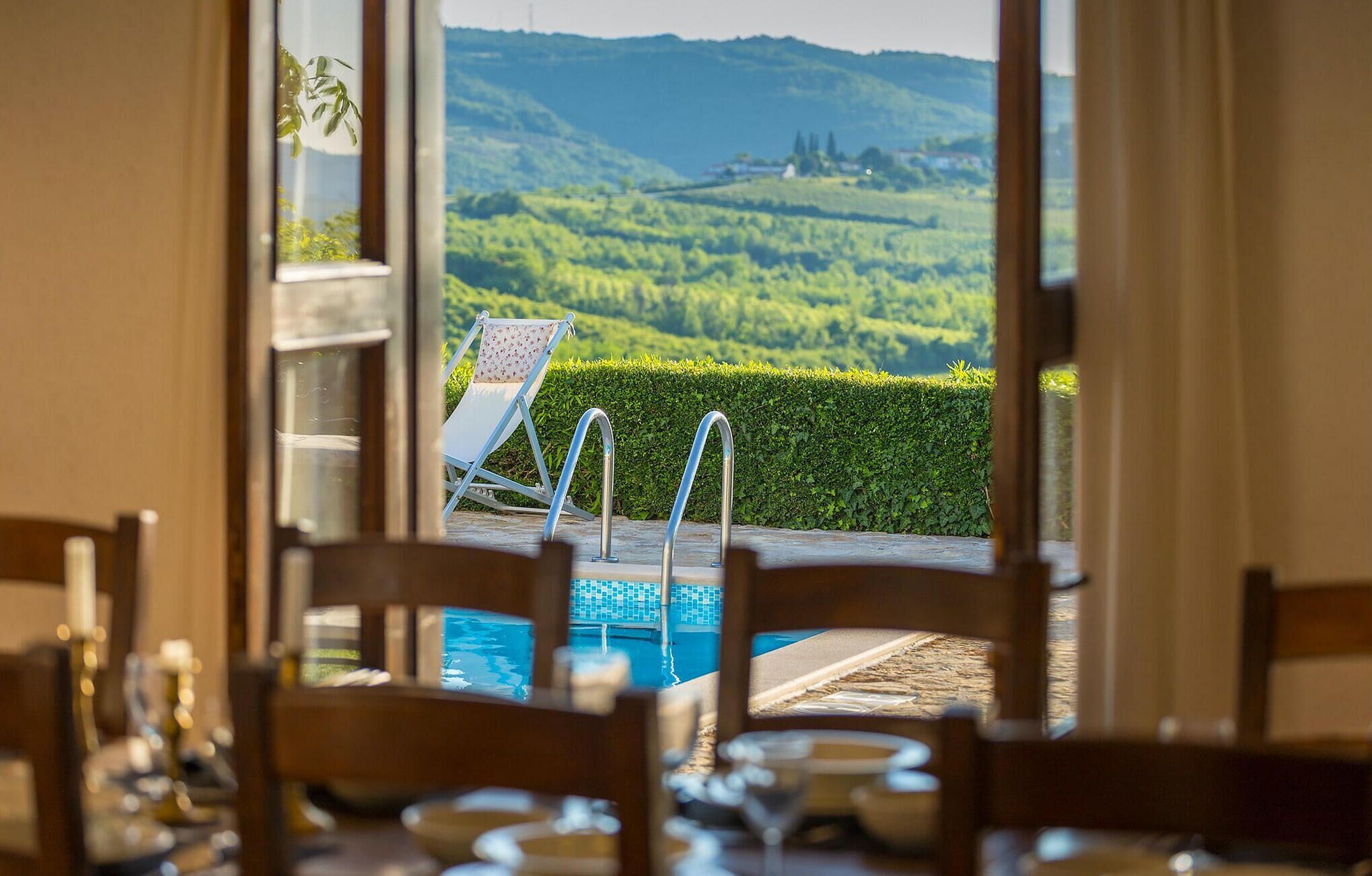 Blick über den Pool mit Stuhl und Tisch, freie Sicht auf das Wasser und eine malerische Landschaft mit Hügeln und einer Hecke.