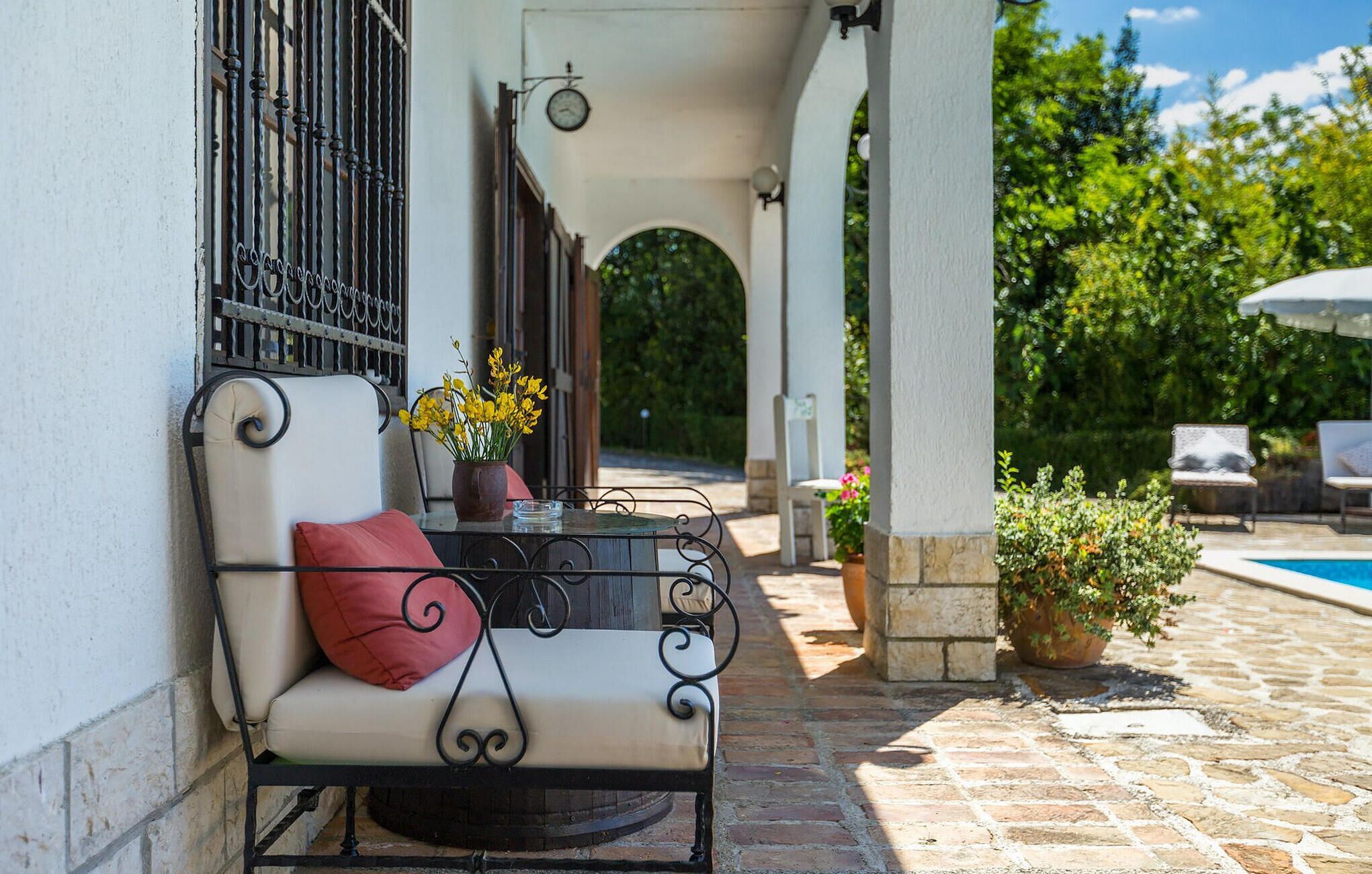 A covered outdoor seating area with a bench, a table with a vase of flowers, and a view of a swimming pool.