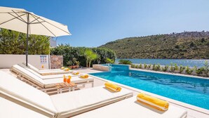 A poolside area with lounge chairs, a sun umbrella, and a view of a lake and hills. - Lovely home in Rogoznica (Rogoznica)
