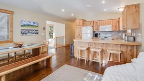 Une cuisine spacieuse avec des meubles en bois et un îlot central, un coin repas avec un banc et une table, et une vue sur l’extérieur par une fenêtre.