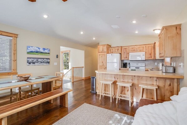 A spacious kitchen with wooden cabinets and a central island, a dining area with a bench and table, and a view of the outdoors through a window.