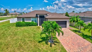 A single-story house with a brick driveway, a front lawn, and a palm tree. - The Cape Escape - New Heated Saltwater Pool.  Just 10 minutes from Matlacha. (Cape Coral)