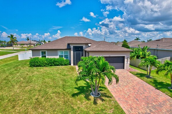 A single-story house with a brick driveway, a front lawn, and a palm tree. - The Cape Escape - New Heated Saltwater Pool. Just 10 minutes from Matlacha. (Cape Coral)