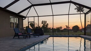 An outdoor swimming pool with lounge chairs and a covered area, surrounded by a fence and palm trees, with a sunset view. - The Cape Escape - New Heated Saltwater Pool.  Just 10 minutes from Matlacha. (Cape Coral)