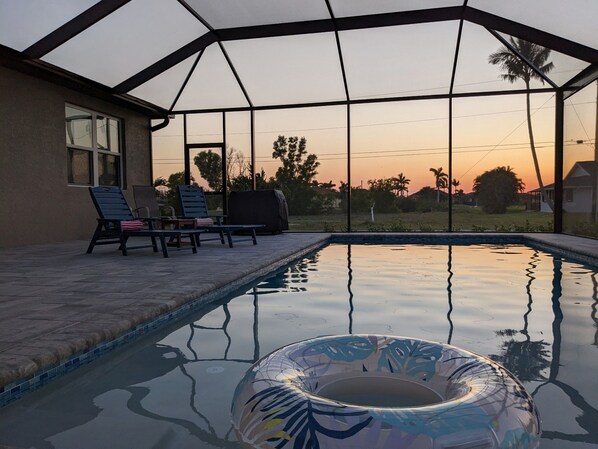An outdoor swimming pool with lounge chairs and a covered area, surrounded by a fence and palm trees, with a sunset view. - The Cape Escape - New Heated Saltwater Pool. Just 10 minutes from Matlacha. (Cape Coral)
