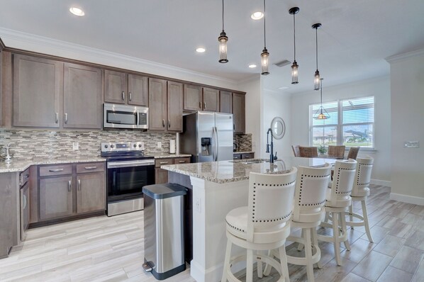 A modern kitchen with wooden cabinets, stainless steel appliances, and a central island with bar stools. - The Cape Escape - New Heated Saltwater Pool. Just 10 minutes from Matlacha. (Cape Coral)