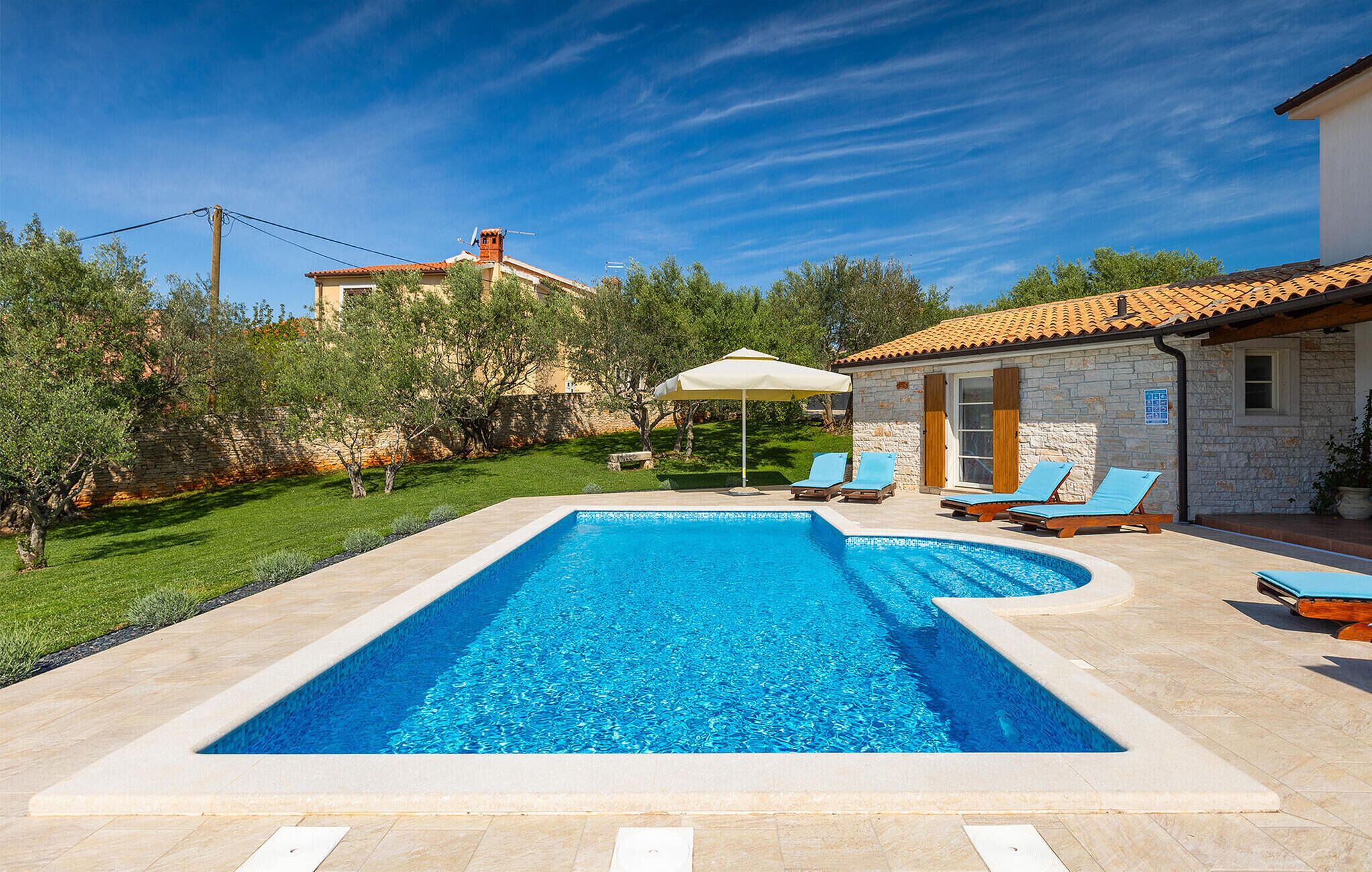 A rectangular swimming pool with clear blue water, surrounded by a stone deck. There are lounge chairs and a sun umbrella near the pool.