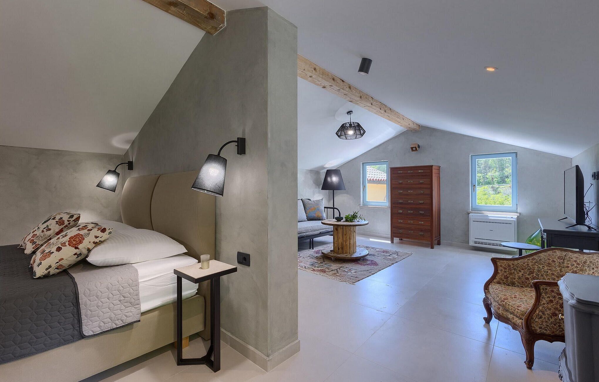 A modern loft bedroom with a sloped ceiling, a large bed with patterned pillows, a bedside table with a lamp, and a view of a dining area with a table and chairs.