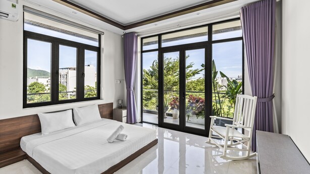 A modern hotel room with a large bed, a rocking chair, and a balcony with a view of greenery and buildings. - Rosemary Villa near Han River  (Da Nang)