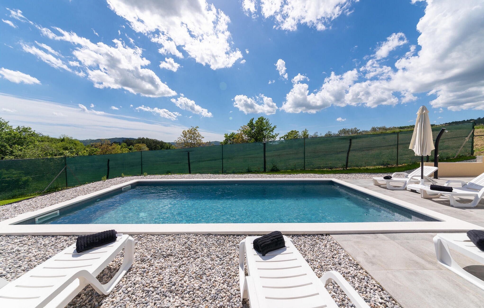 Ein rechteckiger Swimmingpool mit weißen Liegestühlen und einem grünen Zaun im Hintergrund.