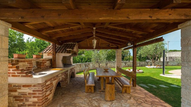 Un patio techado al aire libre con zona de barbacoa de ladrillo, piso de piedra, bancos de madera y pared de piedra.