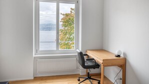 A room with a wooden desk, a black office chair, and a view of a body of water through a window. - Chic Horgen 2BR w/ Storage Room & Lake View, nr Market, by Blueground (Zurich)