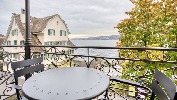 A balcony with a table and chairs overlooking a body of water and buildings. - Chic Horgen 2BR w/ Storage Room & Lake View, nr Market, by Blueground (Zurich)