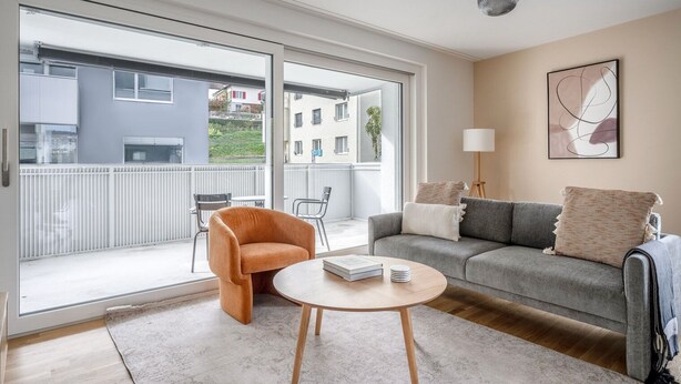 Ett modernt vardagsrum med en grå soffa, en orange fåtölj och ett runt soffbord. Det finns en skjutdörr i glas som leder ut till en balkong.